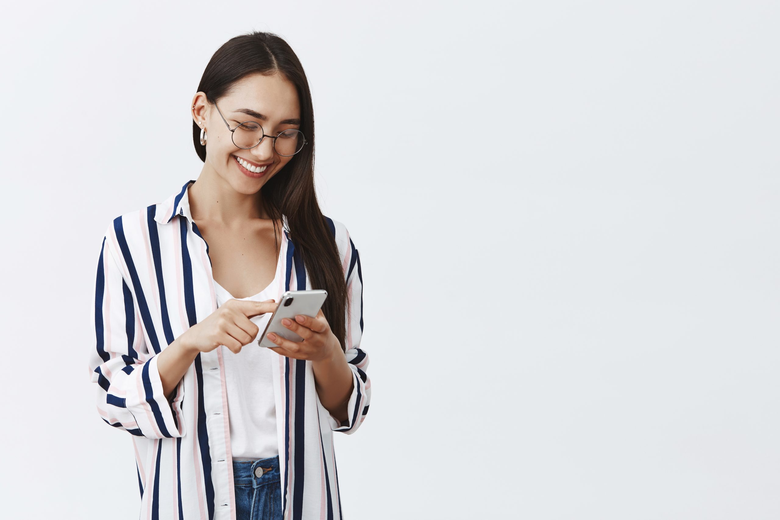 Choosing new wallpaper on smartphone. Portrait of happy attractive and stylish woman in glasses and striped blouse scrolling feed while using smartphone, reading amusing article in internet. Technology concept